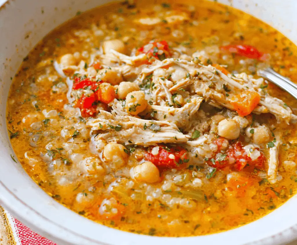 A steaming bowl of hearty chicken and chickpea soup garnished with fresh herbs, showcasing tender chicken pieces, chickpeas, vegetables, and a flavorful broth.